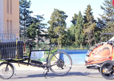 un velo cargo de marque Omnium avec une remorque rouge derrière transportant une grosse caisse noire