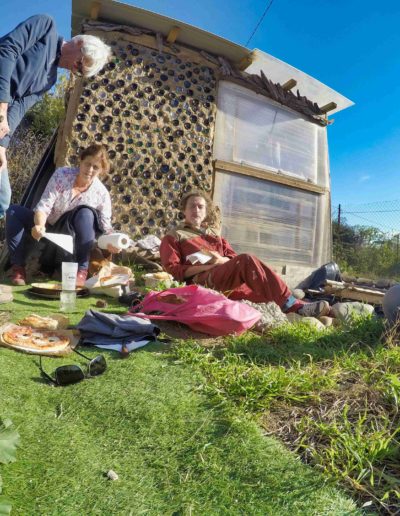 des gens savourant une pause déjeuner devant une serre bioclimatique sous un beau ciel bleu et ensoleillé, dans l'herbe au pied d'un mur en bouteilles de verre.